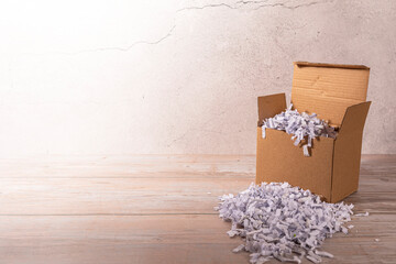 Konzept eines Kartons auf einem Holzboden vor einer weißen Steinwand gefüllt mit geschredertem Papier mit Platz für Text