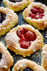 Sweet yeast buns with the addition of berry fruits and butter crumble, sprinkled with powder sugar on a black background, focus on the roll with strawberries, close up view