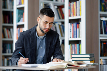 Student wearing eyeglasses writing homework in library, speaking aloud his ideas, heap of books...