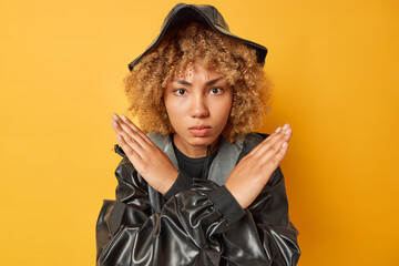 Serious strict woman shows taboo stop gesture crosses hands rejects something disagrees with you prohibits action wears black hat and raincoat isolated over yellow background. Body language concept