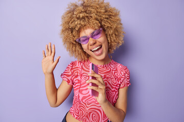 Waist up shot of happy overjoyed woman has curly hair keeps palm raised holds phone near mouth sings song wears purple sunglasses pink tshirt poses indoor has fun. Carefree optimistic female model