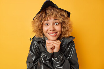 Portrait of cheerful surprised woman with curly hair keeps hands under chin bites lips cannot believe in something unexpected wears black leather jacket and hat isolated over yellow background