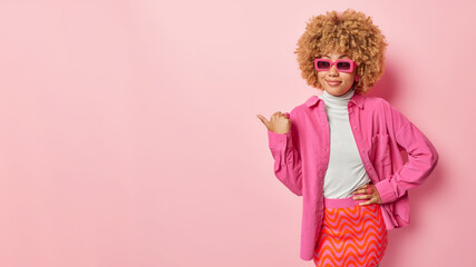Horizontal shot of pleased fashionable curly woman points thumb aside shows place for advert or...