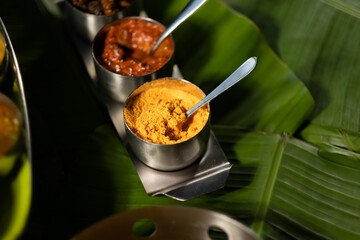 An assortment of traditional Indian seasonings — curry chilis; pickles; and 'gunpowder' splice blend ('milagai podi') — against a backdrop of banana leaves, suggesting southern Indian cuisine.