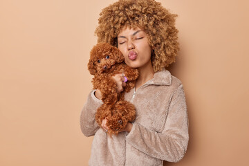 Curly haired young woman poses with cute puppy keeps lips folded dressed in fur jacket poses with affectionate poodle dog isolated over brown background. Domestic animal owner poses indoors.