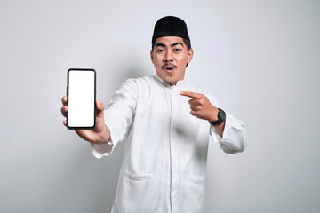 Surprised young Asian Muslim man pointing finger at display mobile phone screen isolated on white background.