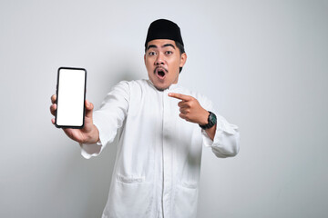 Surprised young Asian Muslim man pointing finger at display mobile phone screen isolated on white background.