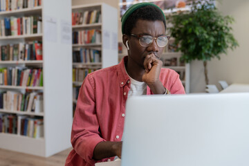 Focused pensive African American hipster freelancer man in eyeglasses working at laptop at library workplace table thinking over IT online startup project. Concentrated black guy student solves task. - obrazy, fototapety, plakaty