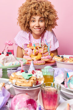 Vertical shot of curly haired woman bites lips surrounded by appetizing delicious creamy desserts celebrates festive occasion comes on party isolated over pink background. Birthday celebration concept