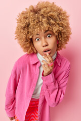 Fototapeta na wymiar Vertical shot of wondered woman with curly hair keeps mouth opened from shock looks curious wears stylish shirt and skirt has widely opened eyes poses against pink background. Amazement concept
