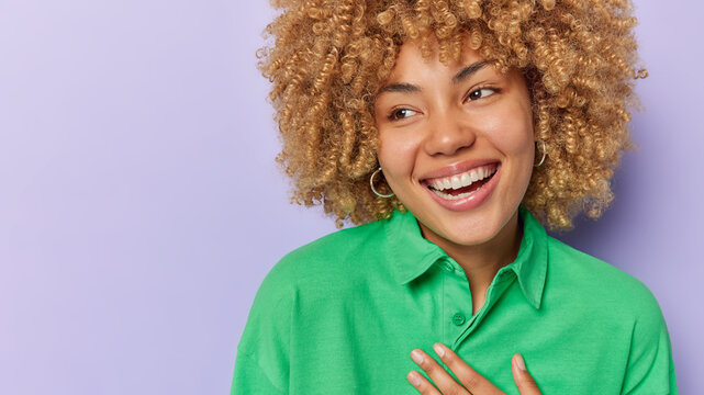 Close up portrait of cheerful good looking woman with blonde curly hair keeps hand on chin smiles sincerely wears green shirt isolated over purple background copy space for your advertising content
