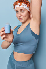Vertical shot of sporty sweaty woman with short red hair applies deodorant under armpit dressed in...