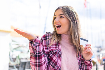 Young pretty Romanian woman holding a credit card at outdoors with surprise facial expression