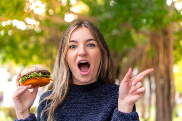 Young pretty Romanian woman holding a burger at outdoors surprised and pointing finger to the side