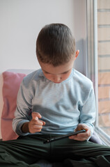 Fototapeta na wymiar Little boy playing mobile game on smartphone sitting on a sofa, top view. Child leisure at home, video gaming addiction