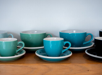 Close-up set of big and small blue green ton of coffee mugs, ceramic cups with saucers stand in a row on wooden shelf isolated on white background with copy space.
