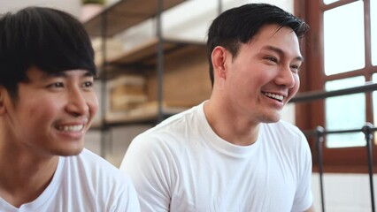 LGBTQ Asian gay couple wearing white t-shirts smiling faces looking happy while watching TV together, gay pride or LGBTQ concept.