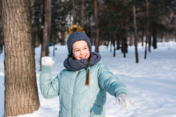 Fototapeta na wymiar A cheerful girl in warm clothes throws a snowball in a winter park. Outdoor games.