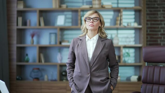 Portrait of serious mature businesswoman in glasses and suit looking at camera while standing in office. Concentrated confident beautiful blonde female. Head shot of a woman at the workplace
