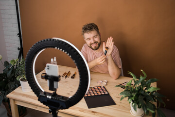 Naklejka na ściany i meble Famous blogger. Cheerful male vlogger showing cosmetics products while recording video and giving advices for his beauty blog. Make-up artist and recording beauty vlog concept