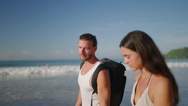 Young biracial happy couple holding hands walking and talking on the beach together enjoying summer closeup. Cheerful boyfriend and girlfriend relaxing and walking by the seaside at sunrise.