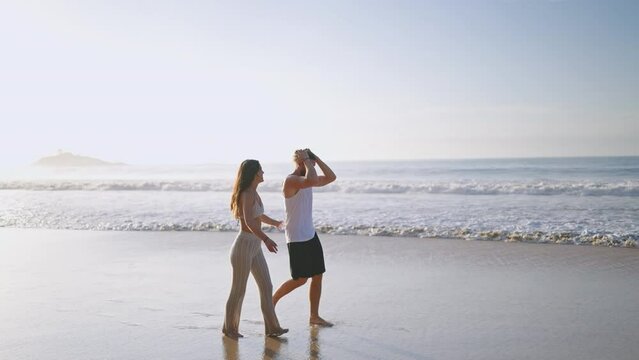 Young biracial happy couple holding hands and walking on the beach together enjoying summer sideview. Cheerful boyfriend and girlfriend relaxing and talking at seaside hugging and kissing at sunrise.