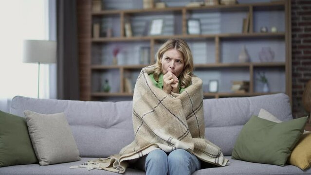 Blonde mature woman is frozen sitting in cold room on sofa at home. A disgruntled female in casual clothes is wrapped in a blanket and warms her hands by rubbing them. Heating problem concept