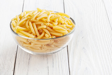 Uncooked maccheroni pasta in glass bowl on white wooden background