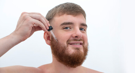 Obraz premium young handsome bearded man apply serum oil, cosmetic product, lotion for healthy perfect fresh skin hydration, grow beard, using skincare, hydrating on white background 