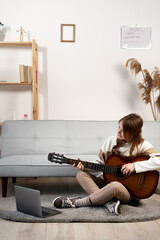 Online music courses. Young woman learns how to play guitar through the internet sitting on the floor at home. Modern technology education benefits concept.