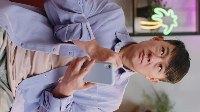 Asian man sits on sofa uses smartphone smiles at home room apartment. Chinese guy texting share messages content on mobile phone social media applications online, watching relax movie. Vertical shot