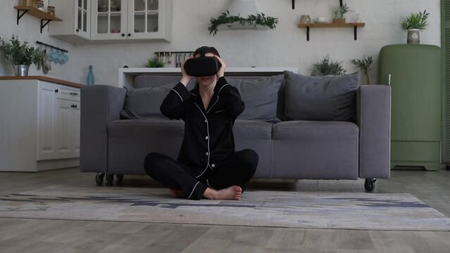 Young woman in pajamas sitting in living room watching video or movie by vr glasses in augmented reality in apartment. Innovation, entertainment and virtual 3D reality, happy woman with futuristic sim