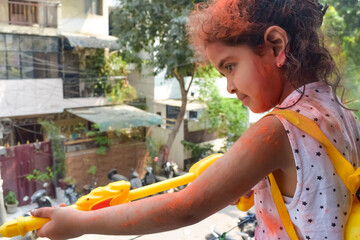 Sweet little Indian girl playing colours on Holi festival, holding pichakaree full of colours, Holi...