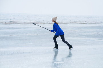 A caucasian woman is skating on a frozen lake holding a selfie stick in her hands. The figure...
