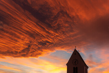 Silhouette of a church steeple under a cloudy orange sky at sunset.