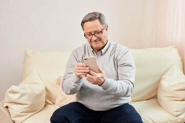 People And Technology. Portrait of smiling mature man using mobile phone, watching video or reading sms message, sitting on the couch in living room at home. Elderly man uses modern technology