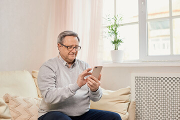 People And Technology. Portrait of smiling mature man using mobile phone, watching video or reading sms message, sitting on the couch in living room at home. Elderly man uses modern technology