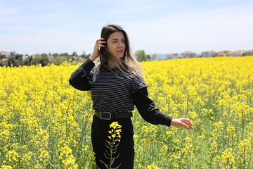 Naklejka na ściany i meble Cute young woman in yellow flower field. Her hair is blowing with wind. She has a black belt around her waist.