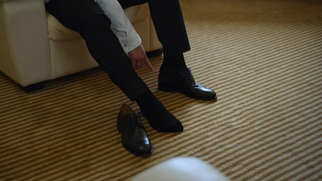 Unrecognizable man in trousers and white shirt on light armchair puts his foot in black leather glossy shoe in room with striped beige carpet. Businessman dressing up for work.