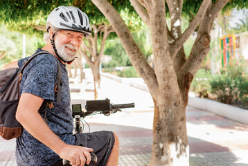 Happy cyclist senior man in urban park wearing helmet and backpack running with electric bicycle.  Concept of healthy lifestyle and sustainable mobility