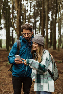 Cheerful millennial caucasian couple in jackets with backpack looking at phone in cold forest, enjoy adventure