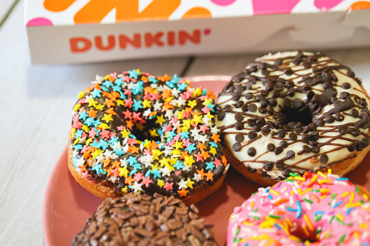 Rosquinhas da marca Dunkin sobre uma t&aacute;bua de madeira e uma caixa de embalagem da marca na composi&ccedil;&atilde;o.	