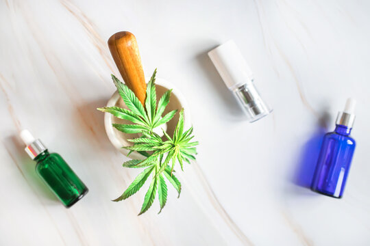 Cosmetic Containers in green, blue and white with dispensers on a marble table. Mortar with a sprout of fresh marijuana on a light background