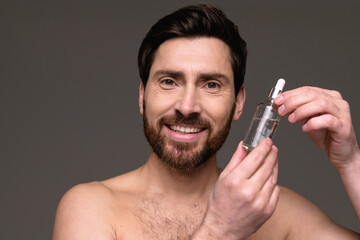 Positive shirtless man holding cosmetic serum isolated on grey background 