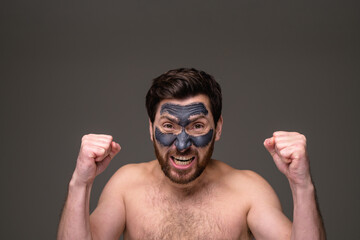 Stressed shirtless man with clay mask on face looking at camera isolated on grey background 