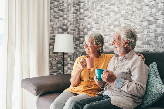 Happy Mature 50s Husband And Wife Sit Rest On Comfortable Sofa In Living Room Enjoy Tea Talking, Smiling Elderly 60s Couple Relax On Couch At Home Drink Coffee Chat Speak Laugh On Leisure Weekend.