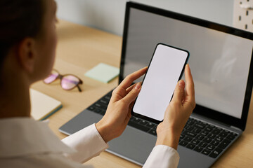 Woman holding smartphone with white screen mockup working at desk