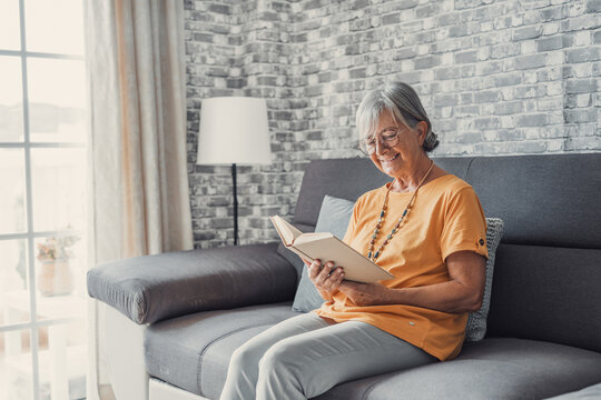 Happy older blonde woman sitting on comfortable sofa in living room, reading interesting book. Smiling elder retired grandmother enjoying leisure weekend time with favorite novel alone at home..