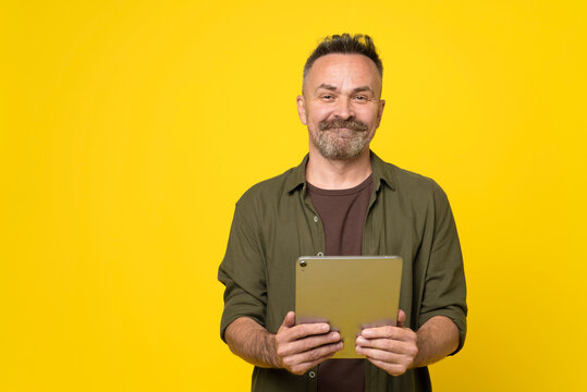 Handsome attractive positive optimist man in green stylish shirt holds tablet pc in hands. Confident middle-aged stubble man looking at camera with tablet PC connected to internet isolated yellow wall