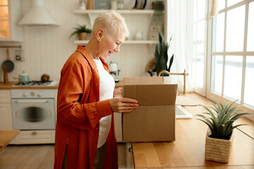 Side view of happy excited stylish mature woman with short haircut standing next to kitchen counter unpacking carton box with goods and products she ordered online. Food delivery service